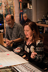 A woman operates a mixing console while a man watches in a recording studio.