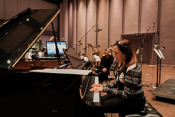 A woman wearing headphones plays a black grand piano in a large recording studio.
