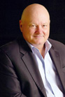 A bald man wearing a dark blazer and a light blue button-down shirt against a black background.