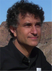 A man with dark curly hair and a black shirt smiling against a rocky mountain background.