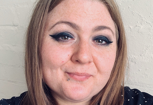 Close-up of a woman with light hair, dark smoky eye makeup, and a small silver lip piercing.