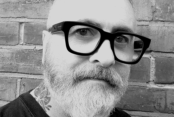 Black and white close-up of a man with thick black glasses and a full grey beard against a brick wall.