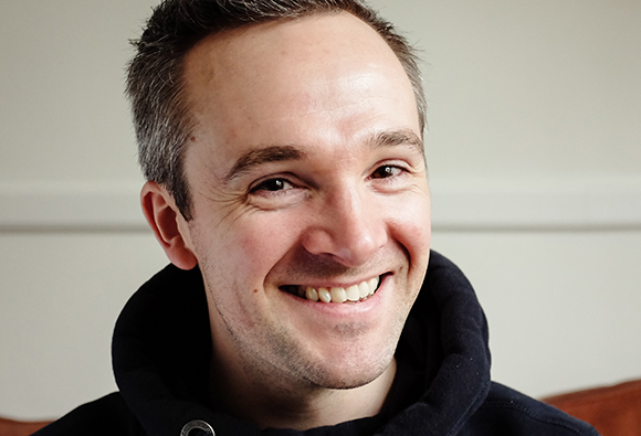 A man with short brown hair smiling broadly against a plain light background.