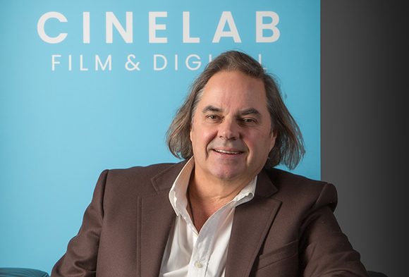 A man with long brown hair wearing a white shirt and brown blazer sits before a blue Cinelab sign.