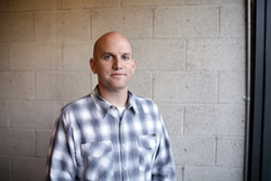 A bald man wearing a grey and white plaid shirt stands before a light grey cinder block wall.