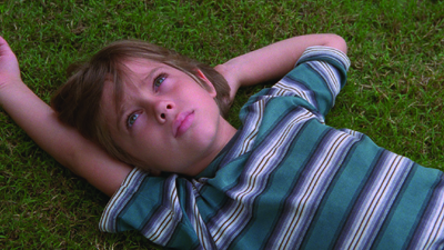 A young child with light hair lies on green grass with hands behind their head, looking upward.