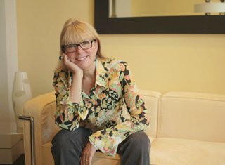A blonde woman wearing glasses and a floral patterned shirt sits on a cream-colored sofa.