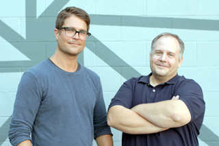 Two men pose together in front of a light blue wall with dark geometric lines.