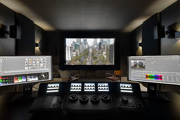 A dark professional post-production studio with dual monitors, a large projection screen, and a grading console.