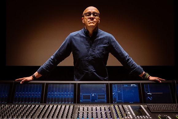 A man in a blue shirt stands behind a large professional audio mixing console with multiple digital screens.