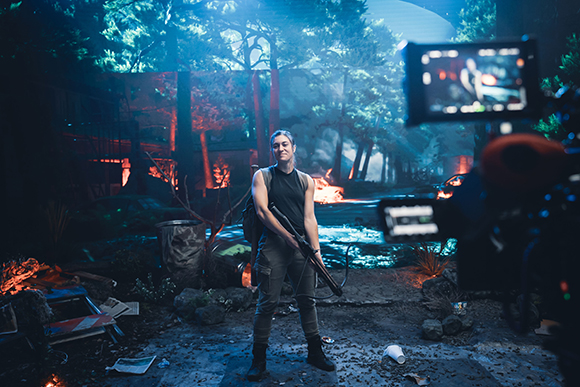 A woman in a black tank top holds a prop rifle on a cinematic forest set with artificial fire.
