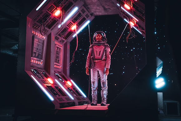 A person in a silver spacesuit stands inside a glowing, hexagonal sci-fi structure against a starry background.