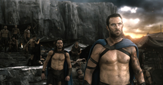 Two shirtless, muscular men in blue capes stand in a rocky camp under a dramatic sunset sky.
