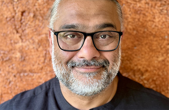 A man with grey hair, a salt-and-pepper beard, and black-rimmed glasses smiling against a textured orange background.