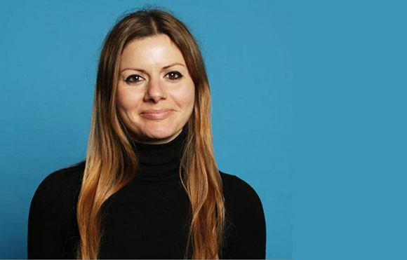 A woman with long brown hair and a black turtleneck smiling against a solid blue background.