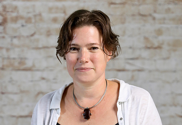 A woman with wavy brown hair and a dark pendant necklace smiles slightly against a light brick wall.