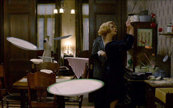 A blonde woman in a dark dress stands in a kitchen while several white plates float mid-air around her.