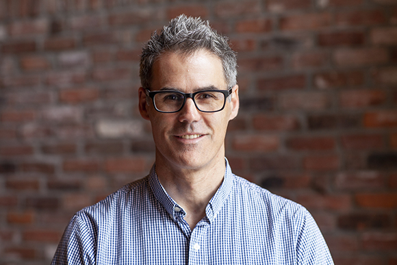 A man with salt-and-pepper hair and black glasses wearing a blue checkered shirt, smiling against a brick wall.