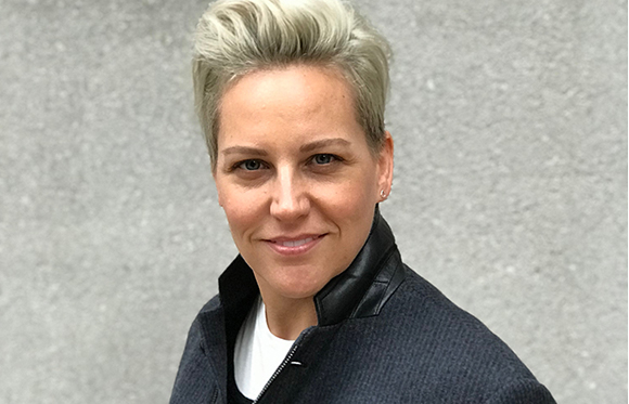 A person with short, styled blonde hair and a slight smile, wearing a dark jacket over a white shirt.