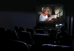 Silhouetted audience members watching a person on a large movie screen in a dark theater.