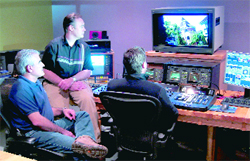 Three men observe video monitors and control panels in a professional editing suite.