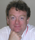 A man with curly brown hair wearing a white shirt, resting his chin on his hand.