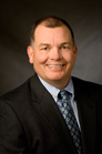 A smiling man with short dark hair wearing a black suit, light blue shirt, and patterned blue tie.