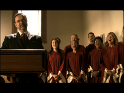 A man in a suit speaks at a wooden podium while a choir in red robes sings behind him.