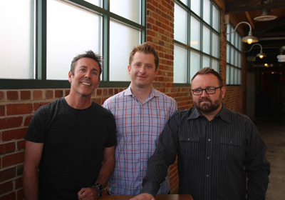 Three men smiling and posing together in an indoor setting with brick walls and large windows.