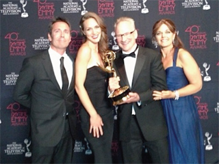 Four people in formal attire pose together holding a gold Emmy award in front of a branded backdrop.