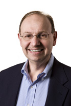 A smiling man with glasses, a light blue button-down shirt, and a dark blazer against a white background.