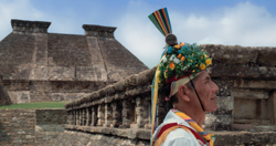 A man wearing a colorful floral headdress and striped ribbons stands in profile before a stone pyramid.
