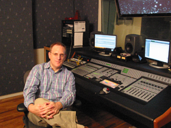 A man in a plaid shirt sits in a chair beside a large professional audio mixing console and computer monitors.