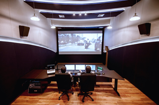 Two people sit at a desk with multiple monitors in a dark control room facing a large projection screen.