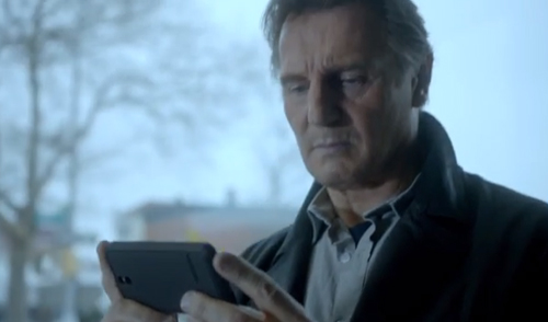 An older man in a dark coat looks down intently at a smartphone held in his hands.