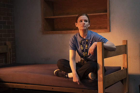A woman wearing a blue polo with an FBI lanyard sits cross-legged on a bed in a dark room.