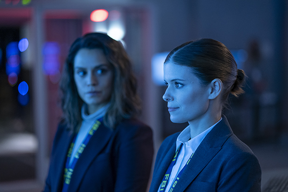 Two women in suits wearing FBI lanyards look intently into the distance in a dark room.