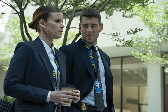 A woman and a man in dark suits wearing FBI lanyards stand together outdoors near trees.