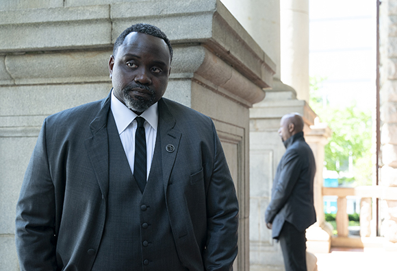 A man with a beard wearing a dark grey suit and tie looks forward against a stone building background.
