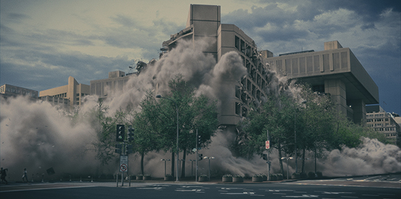 A large concrete building collapses, surrounded by thick, billowing clouds of dust and debris.