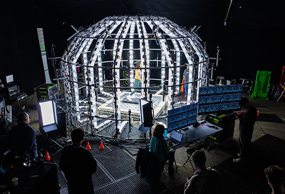 A person stands inside a large, glowing geodesic dome structure surrounded by crew and equipment in a dark studio.