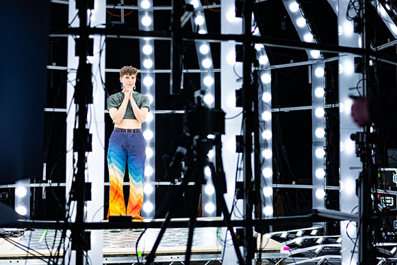 A person in a green crop top and colorful patterned trousers stands on a stage surrounded by studio lights.
