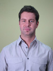 A man with short dark hair wearing a light grey button-down shirt against a plain green background.