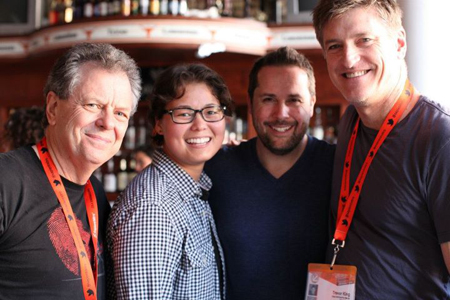 Four people smiling at the camera. Two men on the right wear orange lanyards with badges.