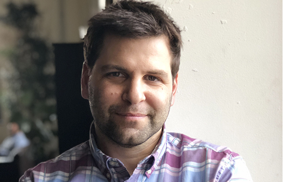 A man with short dark hair and light stubble wearing a purple and blue plaid shirt.