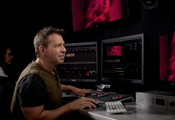 A man sits at a desk using a control panel and mouse to edit video on multiple large monitors.