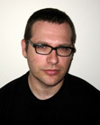 A man with short dark hair and black-rimmed glasses wearing a black shirt against a white background.