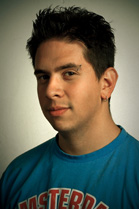 A man with dark, spiked hair wearing a blue t-shirt looks toward the camera against a neutral background.
