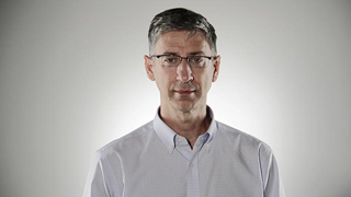 A man with short dark hair and glasses wearing a light blue button-down shirt against a plain background.