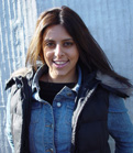A smiling woman with long dark hair wearing a denim jacket and a black puffer vest.